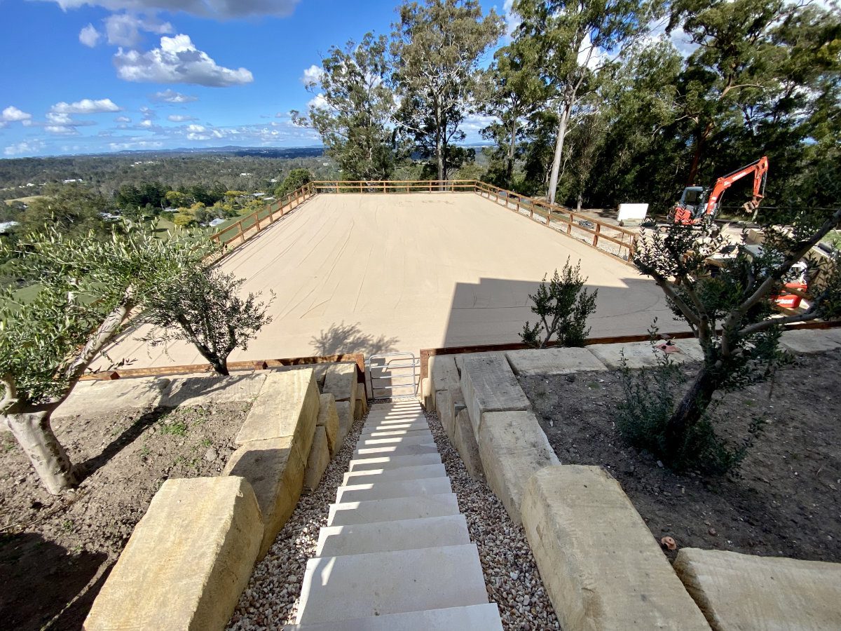 Large, newly constructed sandy court with surrounding trees and a staircase leading to it. Red excavator at a distance.