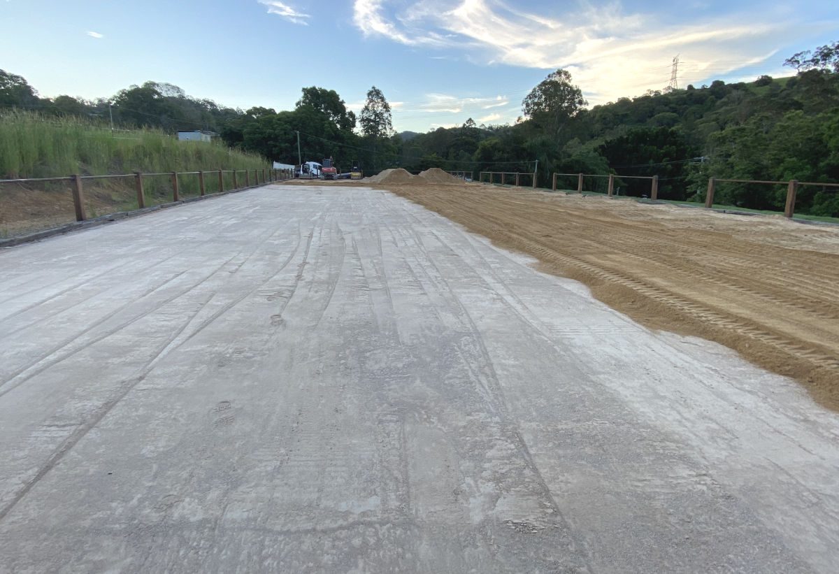 Newly constructed concrete road base with adjacent sandy area and trees under a clear sky Newly constructed concrete road base with adjacent sandy area and trees under a clear sky